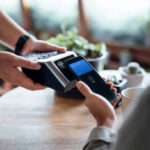 Close up of a male's hand paying bill with credit card contactless payment on smartphone in a cafe, scanning on a card machine. Electronic payment. Banking and technology
